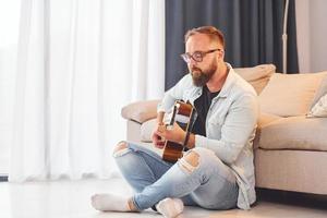 zittend in de buurt de bank. Mens in gewoontjes kleren en met akoestisch gitaar is binnenshuis foto