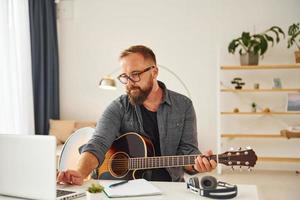 schrijven sommige ideeën. Mens in gewoontjes kleren en met akoestisch gitaar is binnenshuis foto