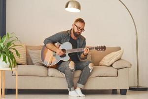 beoefenen liedjes schrijven vaardigheid. Mens in gewoontjes kleren en met akoestisch gitaar is binnenshuis foto