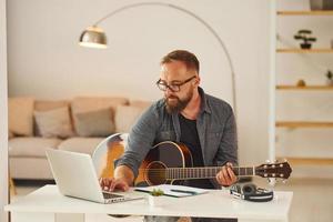 beoefenen liedjes schrijven vaardigheid. Mens in gewoontjes kleren en met akoestisch gitaar is binnenshuis foto