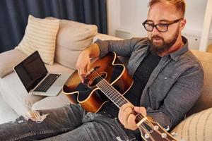 hebben mooi zo tijd beoefenen. Mens in gewoontjes kleren en met akoestisch gitaar is binnenshuis foto