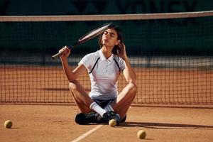 brunette is zittend Aan de grond. vrouw tennis speler is Aan de rechtbank Bij dag foto