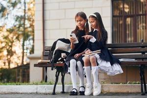 twee schoolmeisjes is zittend buiten samen in de buurt school- gebouw foto