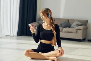 zittend en Holding smartphone. vrouw in sportief kleren aan het doen yoga binnenshuis foto
