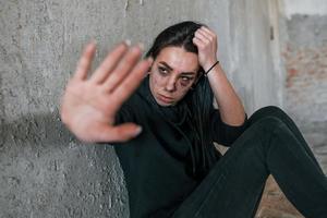 hou op de geweld. jong geslagen vrouw in zwart kleren zittend Aan de verdieping van verlaten gebouw foto