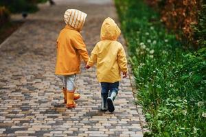 twee kinderen in geel waterbestendig mantels en laarzen houdt elk die van anderen handen en hebben een wandelen buitenshuis na de regen samen foto