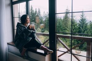 vrouw Holding mok, zittend Aan vensterbank en op zoek Bij bergen foto