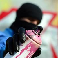 een jong graffiti artiest in een blauw jasje en zwart masker is Holding een kan van verf in voorkant van hem tegen een achtergrond van gekleurde graffiti tekening. straat kunst en vandalisme concept foto