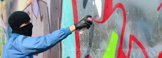 een jong hooligan met een verborgen gezicht verven graffiti Aan een metaal muur. onwettig vandalisme concept foto