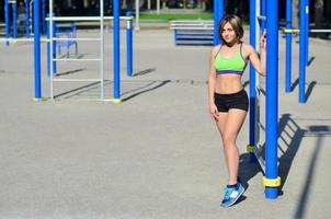 een jong Kaukasisch atleet meisje in een helder groen sportkleding staat tegen de backdrop van een straat sport- speelplaats voor buitenshuis atletiek opleiding. zomer sport- en gezond levensstijl concept foto