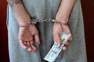 een gearresteerd vent in grijs broek met geboeid handen houdt een reusachtig bedrag van dollar rekeningen. terug visie foto