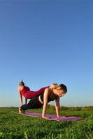 twee jong blond haar meisjes in sport- pakken praktijk yoga Aan een pittoreske groen heuvel in de Open lucht in de avond. de concept van sport oefenen buitenshuis foto