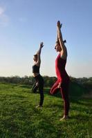 twee jong blond haar meisjes in sport- pakken praktijk yoga Aan een pittoreske groen heuvel in de Open lucht in de avond. de concept van sport oefenen en gezond levensstijlen foto