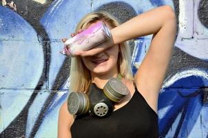 jong en mooi glimlachen sexy meisje graffiti artiest met gas- masker Aan haar nek schuilplaats zijn ogen met een verstuiven kan staand Aan een muur achtergrond met een graffiti patroon in blauw en Purper tonen foto