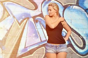 sexy Kaukasisch blond meisje in denim shorts en zwart tank top poseren tegen graffiti muur in de dag buitenshuis foto