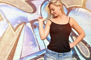 sexy Kaukasisch blond meisje in denim shorts en zwart tank top poseren tegen graffiti muur in de dag buitenshuis foto