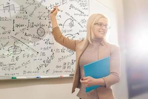 leraar onderwijs hoe naar tellen Aan whiteboard in klas. glimlachen blond vrouw uitleggen toevoegingen in kolom in klas. wiskunde leraar uitleggen rekenkundig bedragen naar elementair kinderen. foto