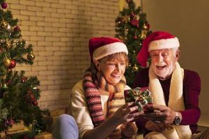 senior Kaukasisch paar vieren Kerstmis samen in geluk en opwinding Bij huis met rood de kerstman hoed en Kerstmis boom foto