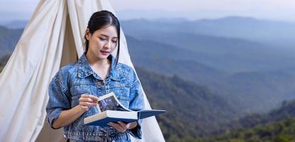 Aziatische vrouw die een boek leest terwijl ze op een solo-trekkingkamp op de top van de berg zit met een kleine tent voor weekendactiviteiten en buitenactiviteiten foto