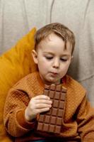 weinig aanbiddelijk jongen zittend Aan de bankstel Bij huis en aan het eten chocola bar. kind en snoepgoed, suiker banketbakkerij. kind genieten een heerlijk nagerecht. peuter- kind met gewoontjes kleding. foto