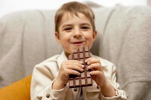 weinig aanbiddelijk jongen zittend Aan de bankstel Bij huis en aan het eten chocola bar. kind en snoepgoed, suiker banketbakkerij. kind genieten een heerlijk nagerecht. peuter- kind met gewoontjes kleding. positief emotie. foto