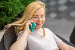 jong blond vrouw zittingen buitenshuis in de stad en maken een telefoontje met haar smartphone. meisje pratend met vrienden met mobiel telefoon. vrije tijd werkzaamheid, communicatie. gesprek. mobiel technologie. foto