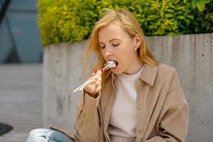 jong mooi blond vrouw aan het eten sushi buitenshuis, Aan de houten terras, door modern gebouw in de stad. smakelijk voedsel naar Gaan. meisje heeft lunch pauze, uitgeven tijd buiten en aan het eten Aziatisch voedsel. stad leven foto