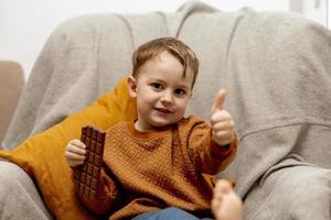 weinig aanbiddelijk jongen zittend Aan de bankstel Bij huis en aan het eten chocola bar. kind en snoepgoed, suiker banketbakkerij. kind genieten een heerlijk nagerecht. peuter- kind met gewoontjes kleding. positief emotie. foto