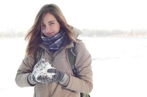 een jong en blij Kaukasisch meisje in een bruin jas houdt een sneeuwbal in de achtergrond van een horizon lijn tussen de lucht en een bevroren meer in winter foto