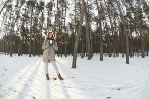 een jong en blij Kaukasisch meisje in een bruin jas houdt een sneeuwbal in een met sneeuw bedekt Woud in winter. vissenoog foto