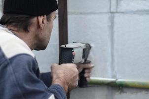 een ouderen werkman boren een gat in een piepschuim muur voor de volgend installatie van een plastic versterken deuvel. creëren gaten in de muur met een oefening. opwarming van de gebouw facade foto