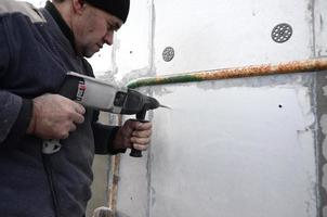 een ouderen werkman boren een gat in een piepschuim muur voor de volgend installatie van een plastic versterken deuvel. creëren gaten in de muur met een oefening. opwarming van de gebouw facade foto