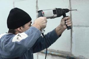 een ouderen werkman boren een gat in een piepschuim muur voor de volgend installatie van een plastic versterken deuvel. creëren gaten in de muur met een oefening. opwarming van de gebouw facade foto