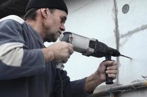 een ouderen werkman boren een gat in een piepschuim muur voor de volgend installatie van een plastic versterken deuvel. creëren gaten in de muur met een oefening. opwarming van de gebouw facade foto