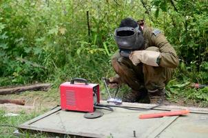 oud Mens lasser in bruin uniform, lassen masker en lassers leer, lassen metaal deur met boog lassen machine buitenshuis foto