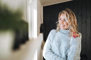 portret van denken glimlachen vrouw veertig jaar met blond gekruld haar- in grijs gebreid trui in de buurt venster Bij huis foto