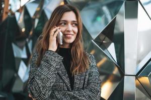 jong glimlachen vrouw in jas met koffie kop gebruik makend van mobiel telefoon in avond stad straat foto