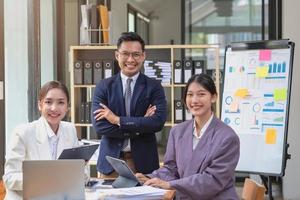 team bedrijf drie glimlachen en gelukkig Aziatisch mannen en Dames team gebruik makend van tablets en laptops brainstorm en verhaal statistieken Aan een zakelijke tabel bord binnen de bedrijf. collega's concept. foto