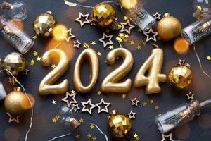 de gouden figuren 2024 gemaakt van kaarsen Aan een zwart steen leisteen achtergrond zijn versierd met een feestelijk decor van sterren, pailletten, Spar takken, ballen en slingers. groet kaart, gelukkig nieuw jaar. foto