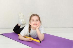 schattig meisje in een sport- uniform leugens Aan een tapijt Holding een gymnastiek- stok in haar handen en glimlacht foto