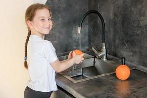 schattig weinig meisje wast een oranje in de wastafel in de keuken en glimlacht foto