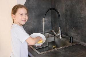 schattig weinig meisje met een varkensstaart wast gerechten in de keuken Bij de wastafel en glimlacht foto