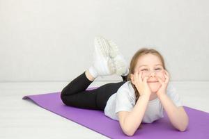 schattig weinig meisje in een sport- uniform leugens Aan een gymnastiek- mat met haar hoofd in haar handen en glimlacht foto