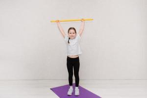 schattig weinig meisje staat Aan een gymnastiek- mat Holding omhoog een gynastiek stok, kinderen geschiktheid foto