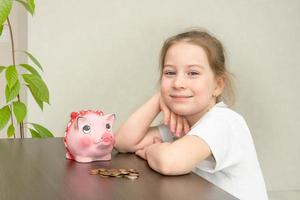 weinig meisje zit Bij een tafel in voorkant van haar met een stack van varkentje bank en verspreide munten, financieel geletterdheid foto