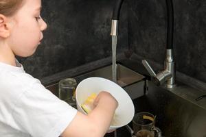 schattig weinig meisje het wassen gerechten in de keuken door de wastafel foto