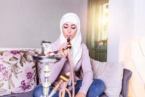 moslim vrouw roken shisha Bij huis. moslim jong vrouw genieten van terwijl roken nargiel. Arabisch meisje roken hookah foto