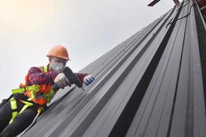 dakdekker arbeider in beschermend uniform slijtage en handschoenen, dakbedekking gereedschap, installeren nieuw daken onder constructie, elektrisch boren gebruikt Aan nieuw daken met metaal vel. foto