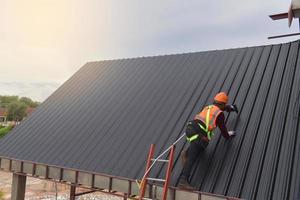 dakdekker arbeider gebruik de ladder in beschermend uniform slijtage en handschoenen, dakbedekking gereedschap, installeren nieuw daken onder constructie, elektrisch boren gebruikt Aan nieuw daken met metaal vel. foto