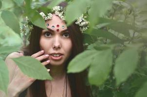 portret van een emotioneel jong meisje met een bloemen krans Aan haar hoofd en glimmend ornamenten Aan haar voorhoofd. schattig brunette poseren in een ontluikend mooi Woud in de dag Aan een prima dag foto